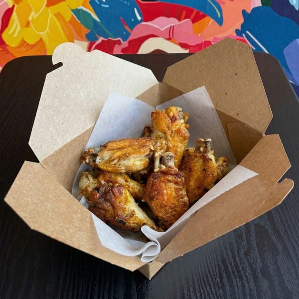Crispy chicken wings in a brown takeout box with white parchment paper, on a dark table.