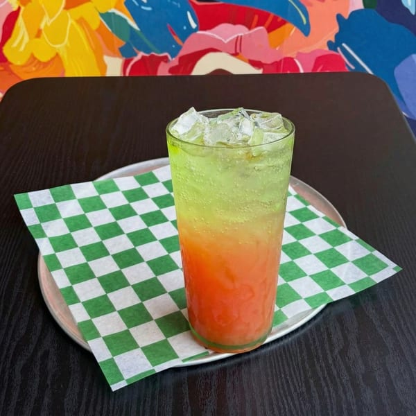 A vibrant green and orange layered drink with ice, on a checkered napkin and metal tray.