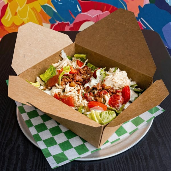 A chicken salad with bacon and tomatoes in a brown takeout box on a checkered paper.