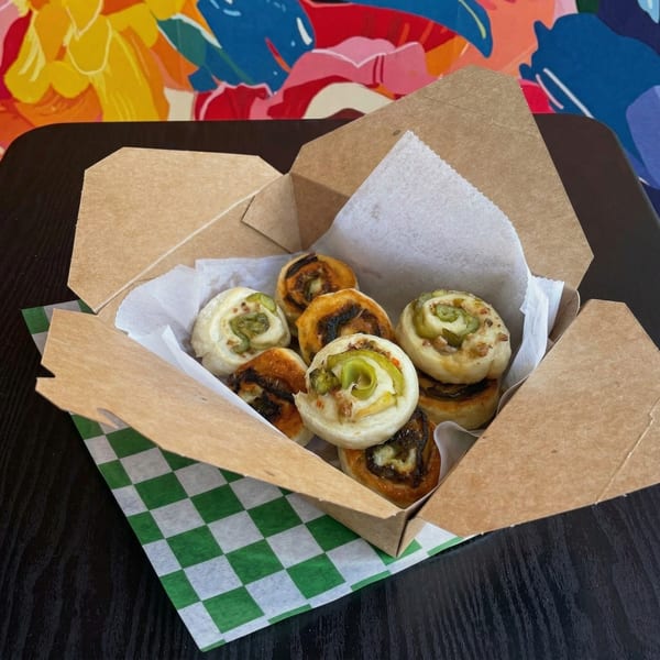 A cardboard takeout box filled with savory pinwheel pastries, some topped with green peppers.
