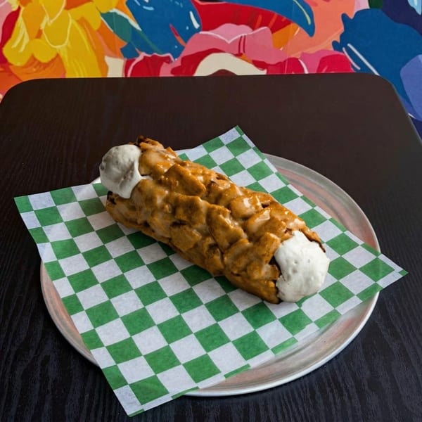 A long, golden-brown dessert with white cream on a green and white checkered paper on a metal plate.