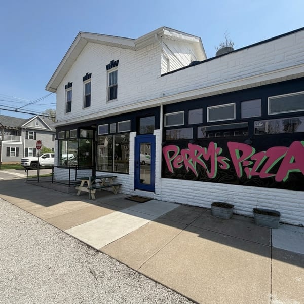 Exterior of Perry's Pizza, a white building with a blue door and pink graffiti-style sign.