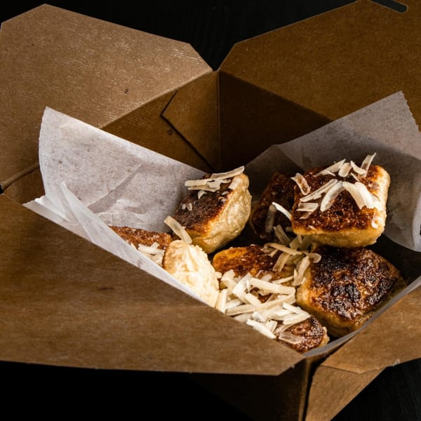 Golden-brown fried plantains topped with shredded coconut, served in a brown takeout box.